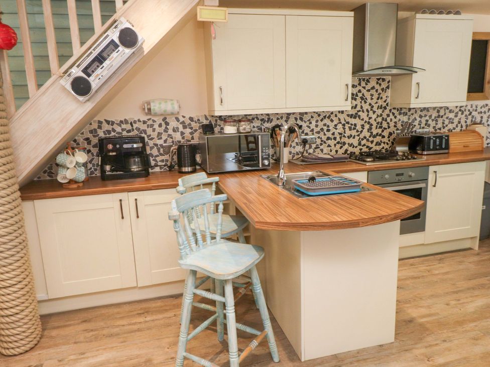 A kitchen with wooden countertops, two light blue bar stools, a coffee maker, microwave, toaster, stove, and a radio under the stairs at Y Stabl in Criccieth