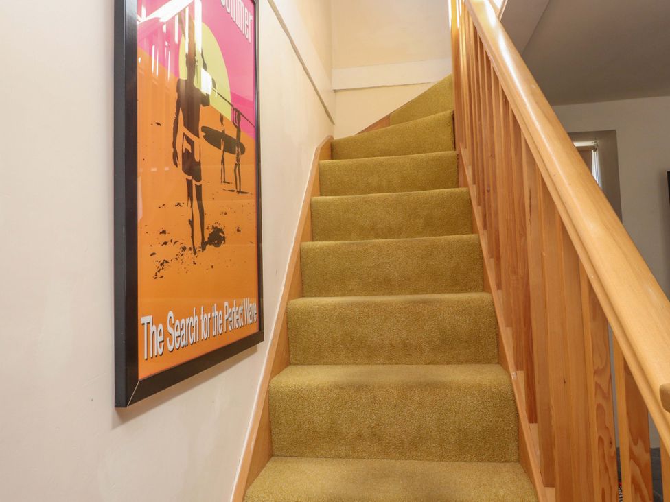 A staircase with yellow carpet and wooden railing next to a surf poster on the wall at Y Stabl in Criccieth