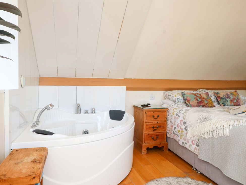 A bedroom with a corner bathtub a wooden nightstand and a bed with patterned bedding at Y Stabl in Criccieth