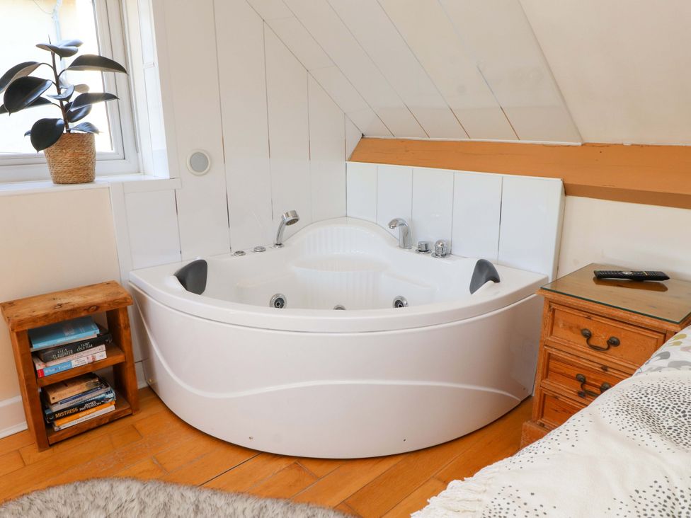 A corner bathtub next to a window with a plant on the windowsill and wooden bedside tables with books and a remote control at Y Stabl in Criccieth