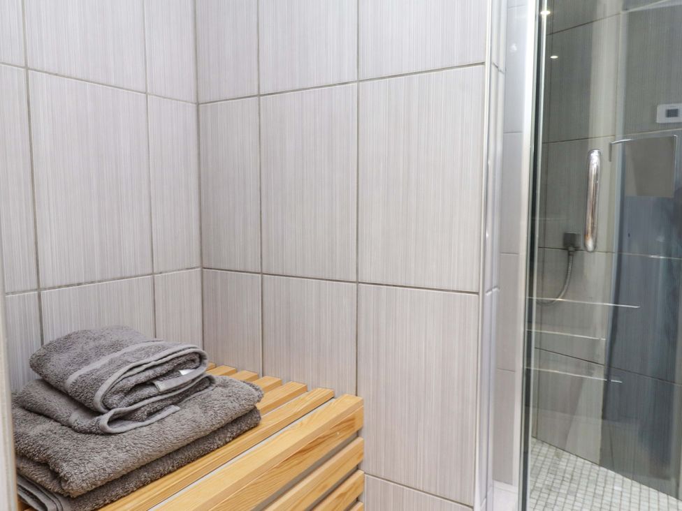 A corner of a bathroom with gray towels on a wooden bench and a glass shower door