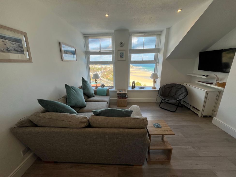 A living room with a sofa and cushions facing two windows overlooking a beach with lamps and a chair next to a mounted TV at Trevose in St Ives