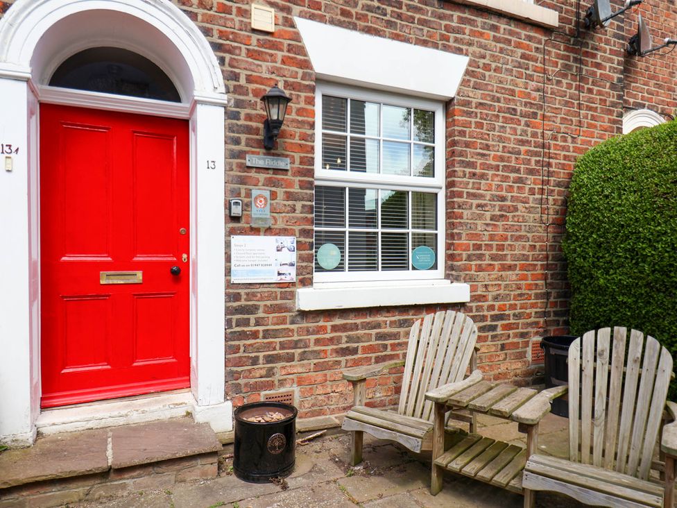 A red door with the number 13 on a brick building next to two wooden chairs and a small wooden table at The Riddle in Whitby