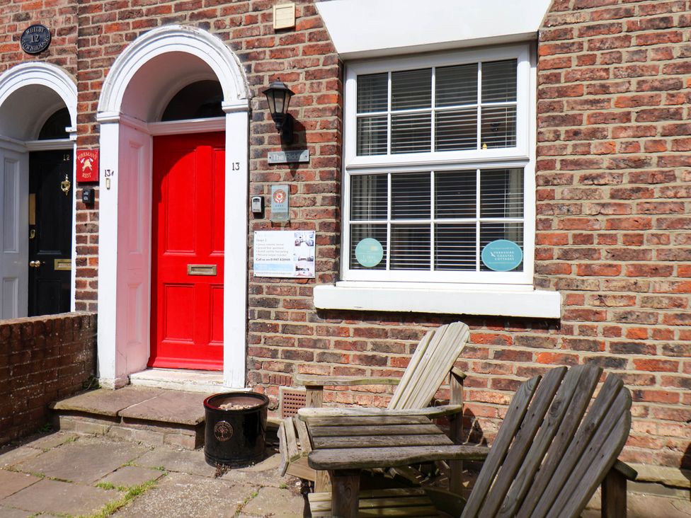 A red front door with two wooden chairs outside a brick house at The Riddle in Whitby