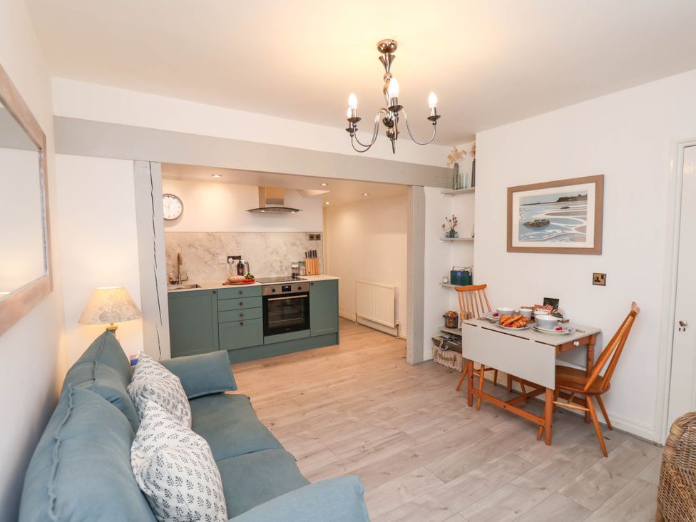 A living area with a blue sofa and a small dining table with two wooden chairs next to an open kitchen with green cabinets at The Riddle in Whitby