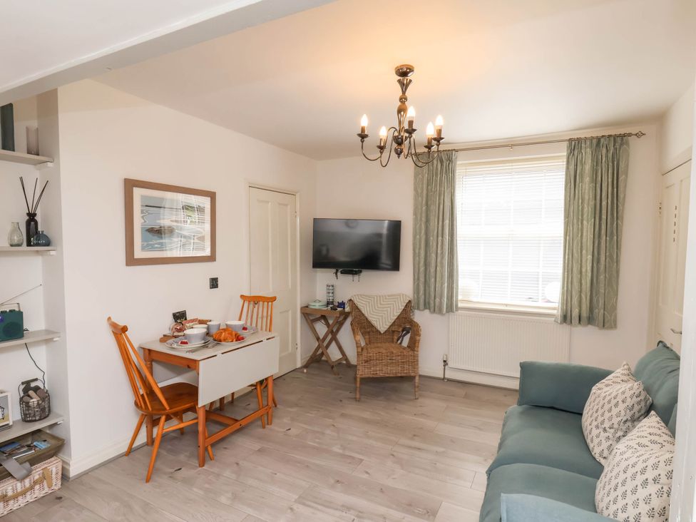 A living room with a dining table and chairs a sofa with cushions a wicker chair a wall mounted TV and a window with curtains at The Riddle in Whitby