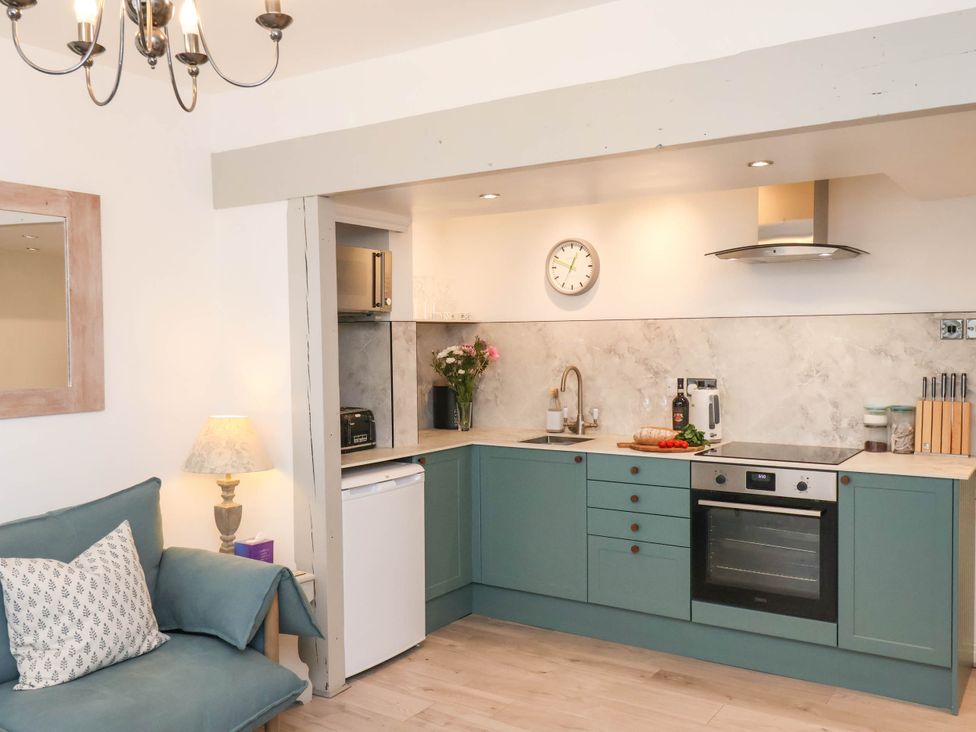 A kitchen with green cabinets, a stove, oven, sink, and a living area with a blue sofa and a lamp at The Riddle in Whitby