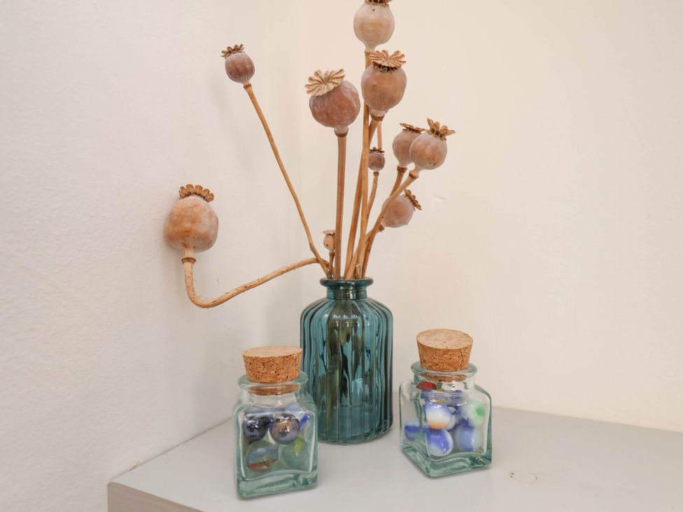 A blue glass vase with dried seed pods and two small glass jars with cork lids containing marbles on a white surface at The Riddle in Whitby