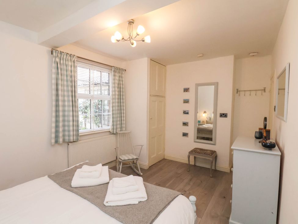 A bedroom with a bed, folded towels, rocking chair, window with curtains, wall mirror, small stool, and white dresser at The Riddle in Whitby