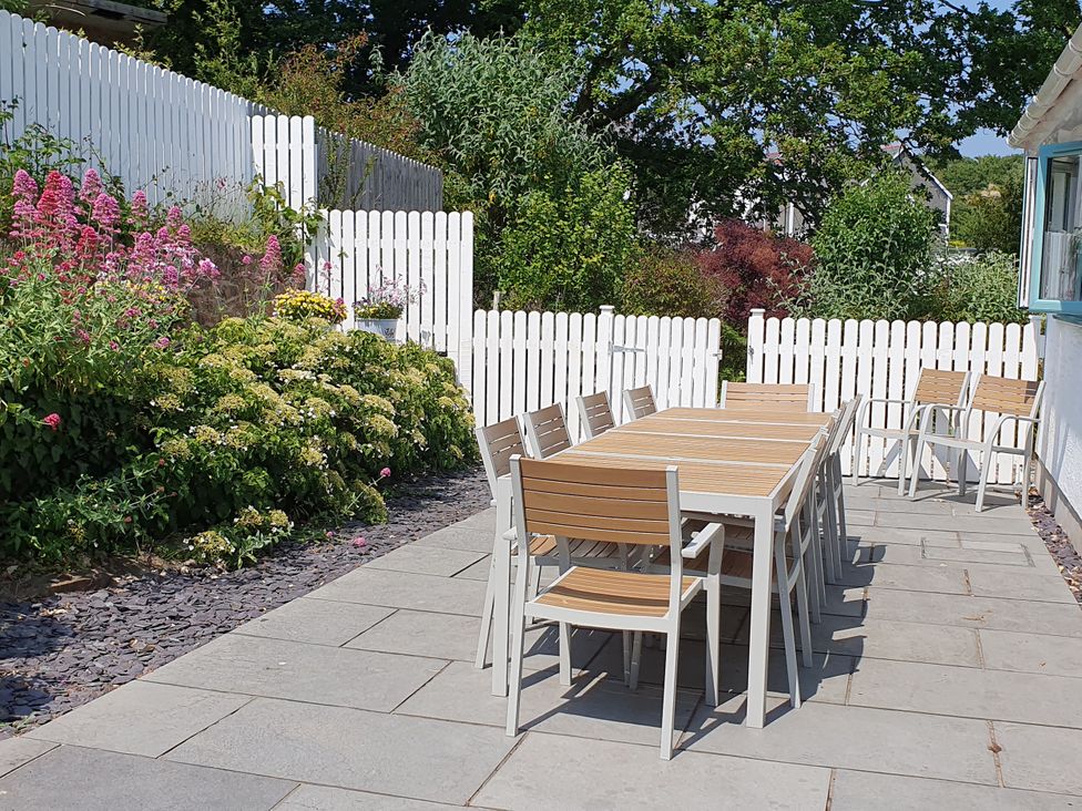 An outdoor patio with a long dining table and chairs next to a white picket fence and garden at Argraig in Llanbedrog