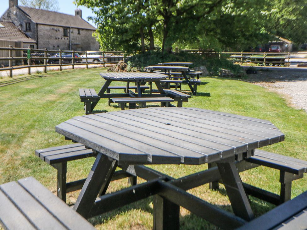 A yard with multiple wooden picnic tables on grass near a stone building and a gravel driveway at Woodlands in Cowdale near Buxton