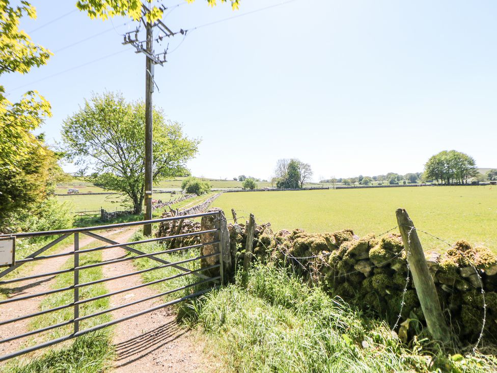A metal gate leading to a dirt path with a telephone pole and stone wall along a green field with trees in the distance at Woodlands in Cowdale near Buxton