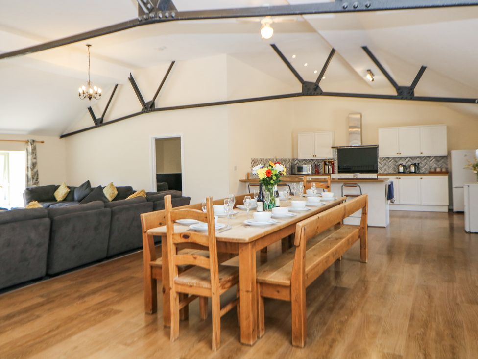 A dining area with a wooden table and chairs set with dishes and glasses next to a kitchen with white cabinets and a living area with sofas at Meadow View in Buxton
