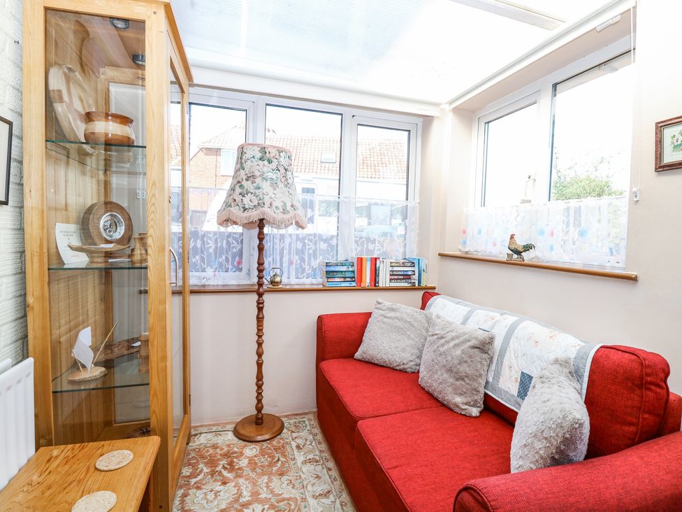 A conservatory with a red sofa and gray cushions a wooden display cabinet with decorative items and a floral floor lamp at Lillies Pad Methwold near Feltwell