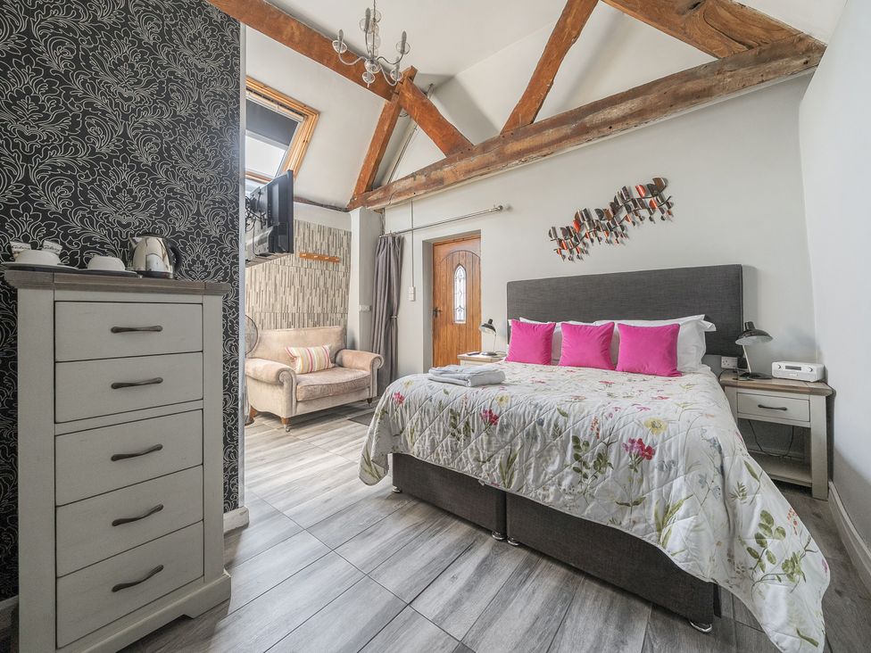 A bedroom with a large bed and pink pillows a dresser and an armchair with exposed wooden beams at The Boars Head Pub Bishop's Castle