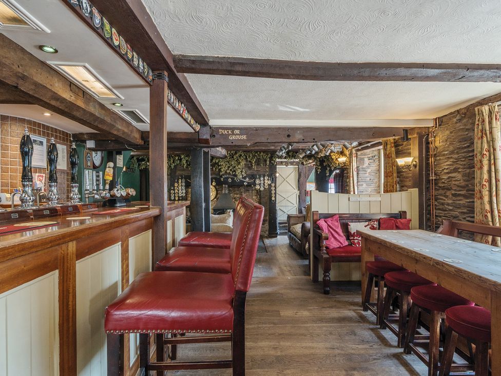 An interior of a pub with a wooden bar counter and red cushioned bar stools a wooden table with red cushioned stools a lounge area with wooden benches and cushions wooden beams on the ceiling and stone walls at The Boars Head Pub in Bishops Castle
