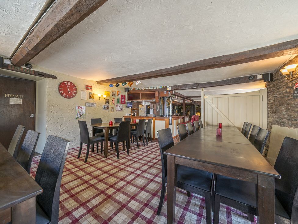 A dining area with multiple wooden tables and chairs a bar counter and a checked carpet at The Boars Head Pub in Bishops Castle