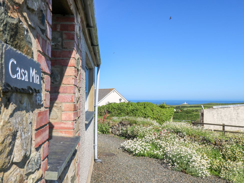 Side view of a stone and brick house with a sign saying Casa Mia and a garden with flowers and greenery and a distant lighthouse and ocean at Casa Mia in Strumble Head Goodwick