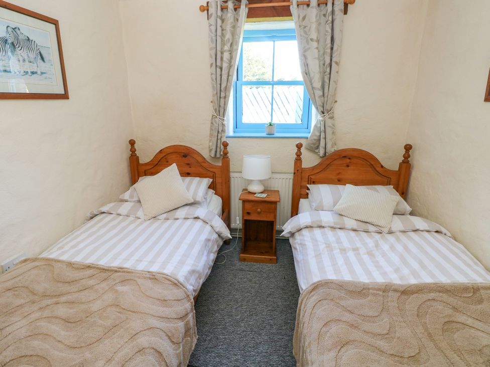 A bedroom with two single beds separated by a wooden nightstand with a lamp and a window with curtains at Casa Mia in Strumble Head Goodwick