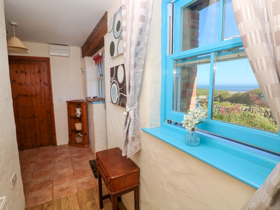 A hallway with a wooden door beige walls a small wooden table and shelves a bright blue window frame with curtains and a vase with white flowers at Casa Mia in Strumble Head Goodwick