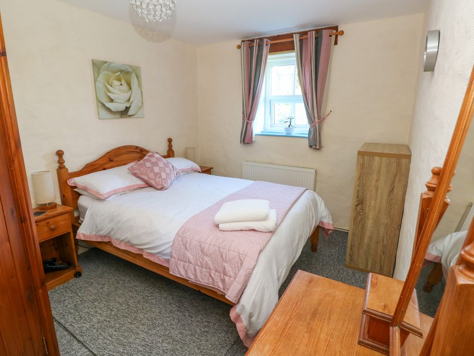 A bedroom with a double bed with pink and white bedding a window with curtains wooden furniture and a flower painting on the wall at Casa Mia in Strumble Head Goodwick