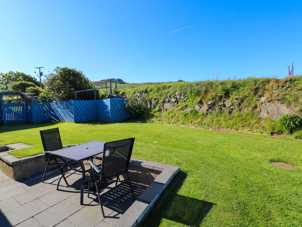 An outdoor patio with a table and two chairs on a stone floor next to a grassy yard with a blue wooden fence at Casa Mia in Strumble Head Goodwick