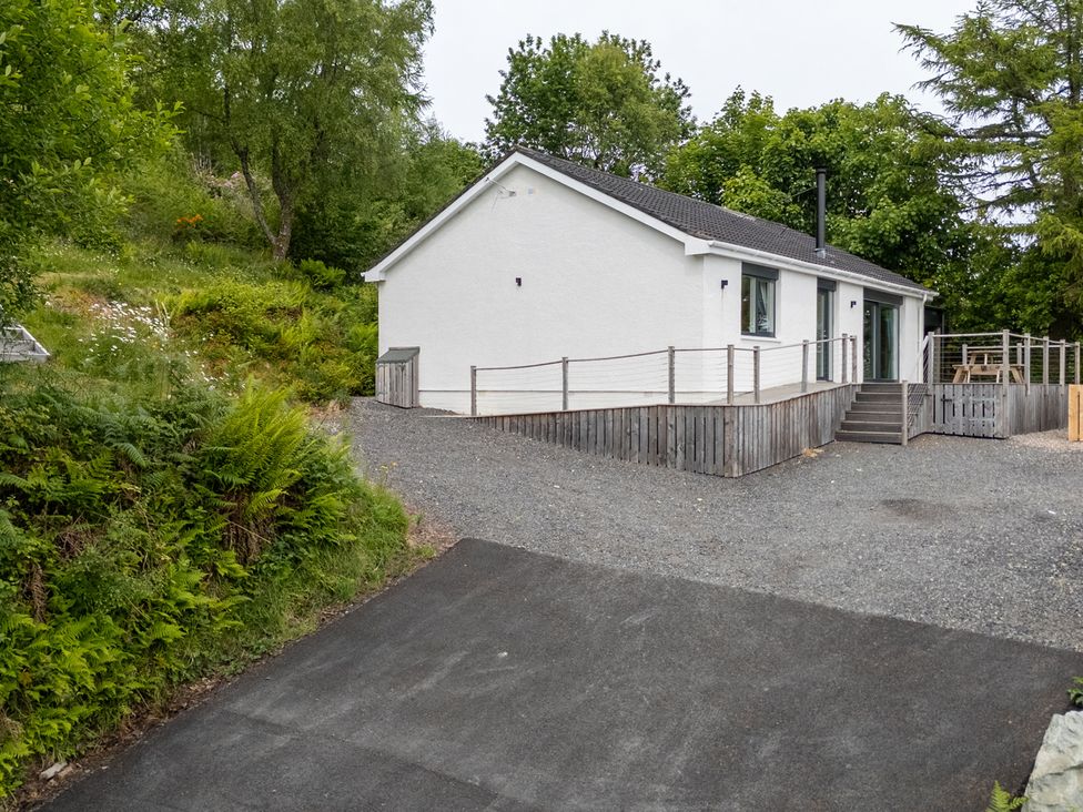 A white single-story house with a wooden deck and gravel driveway surrounded by trees and greenery at The Lookout in Saasaig near Broadford