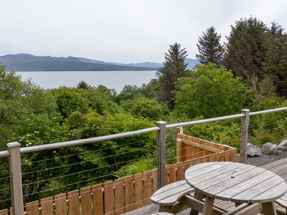 A wooden outdoor table and benches on a deck overlooking trees and a lake with mountains in the background at The Lookout in Saasaig near Broadford