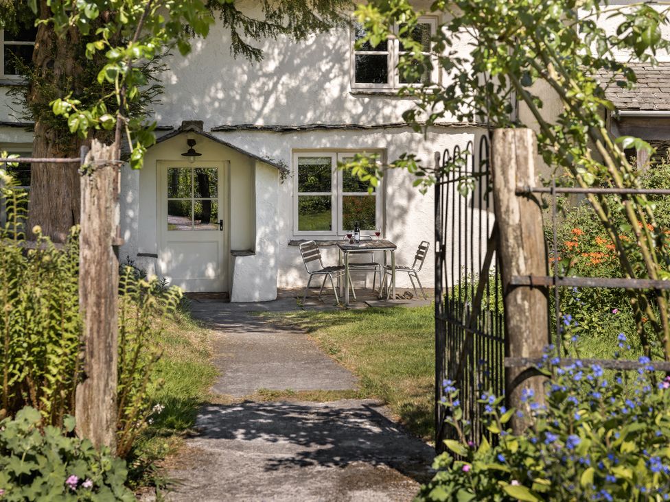 A pathway leading to a white cottage entrance with a table and two chairs outside surrounded by plants and flowers at High Lindeth in Bowness