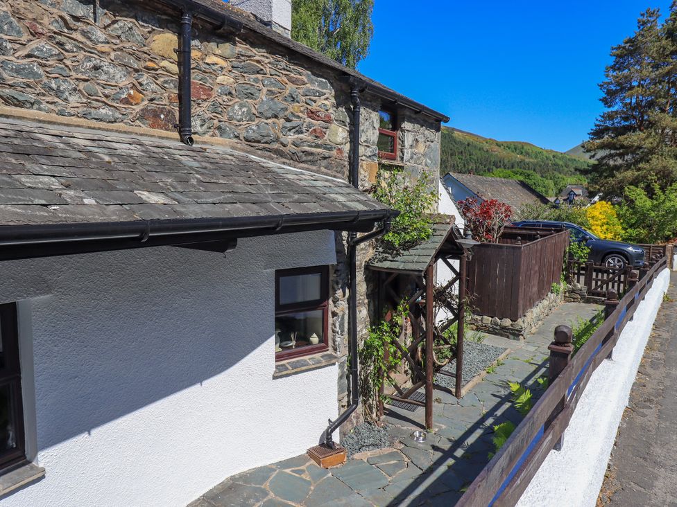 A stone and white plaster house with a slate roof a wooden trellis with climbing plants a paved walkway and a parked car behind a wooden fence at The Cottage in Keswick