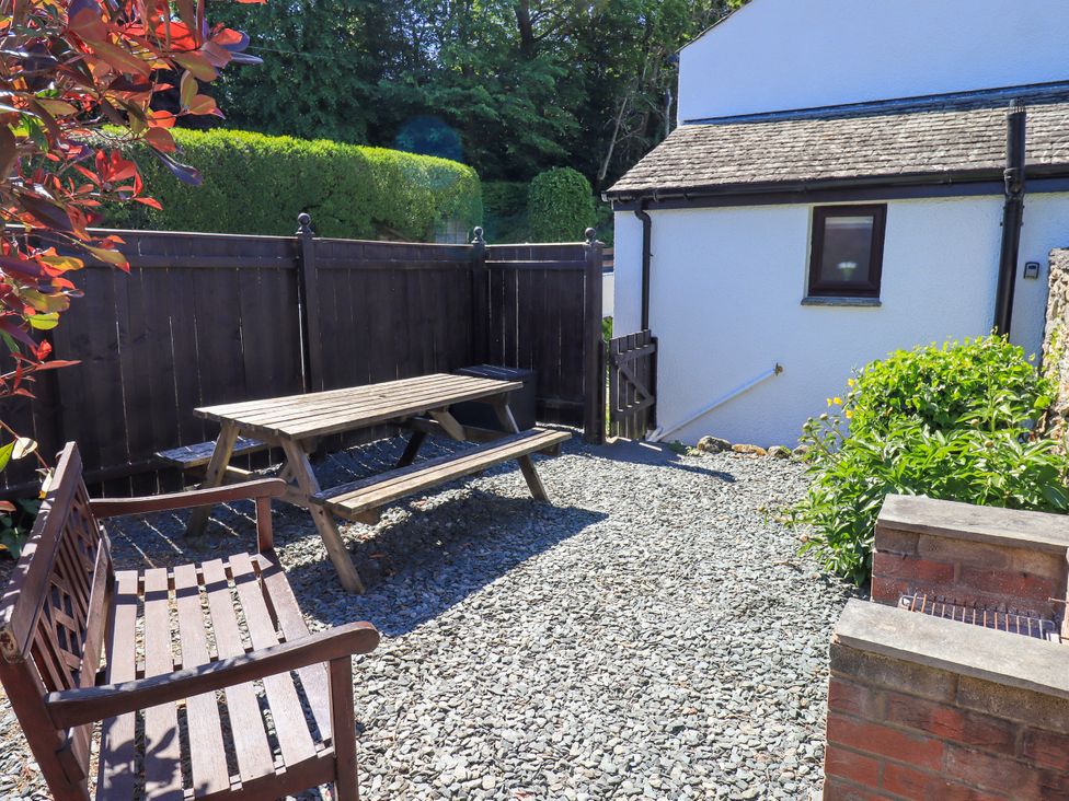 An outdoor patio area with a wooden picnic table and bench next to a white building with a small garden and wooden fence at The Cottage in Keswick