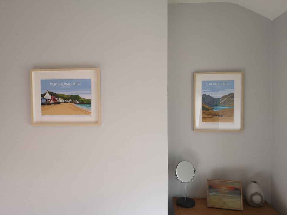 A wall with two framed landscape prints above a wooden surface holding a round mirror a painting and a vase at Y Bwthyn in Llanbedr