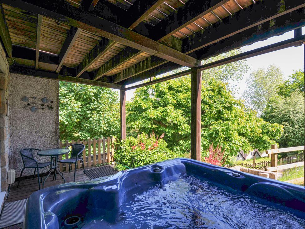A covered outdoor deck with a hot tub and a small table with two chairs surrounded by trees at 59 Valley Lodge in Callington near Gunnislake