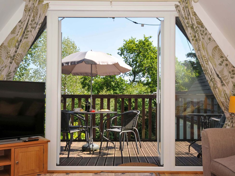 A balcony with a table umbrella and two chairs viewed from a room with a television and curtains at 59 Valley Lodge Callington near Gunnislake