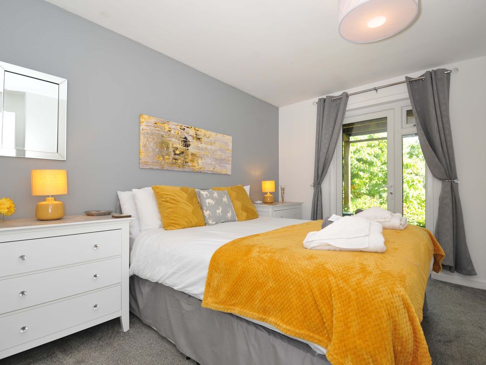A bedroom with a bed with yellow pillows and blanket beside two white drawers with lamps and a window with gray curtains at 59 Valley Lodge Callington near Gunnislake