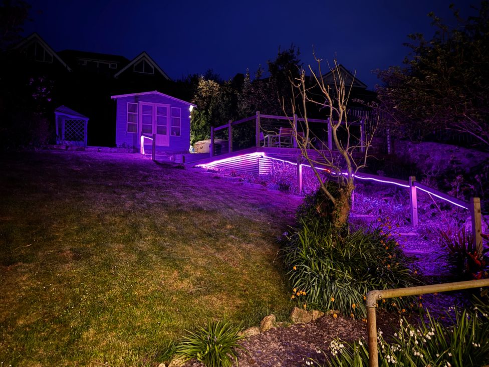 A garden at night with a small shed a patio area with chairs and purple lighting on the railing and steps at Overchurch in Hythe
