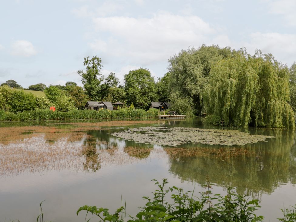 A lake with huts and trees at Willow Lodge At Bridge Lake Farm & Fishery Chacombe
