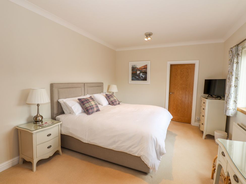 A bedroom with a bed pillows bedside tables lamps a dresser with a television a door and a window at Edencroft in Greystoke