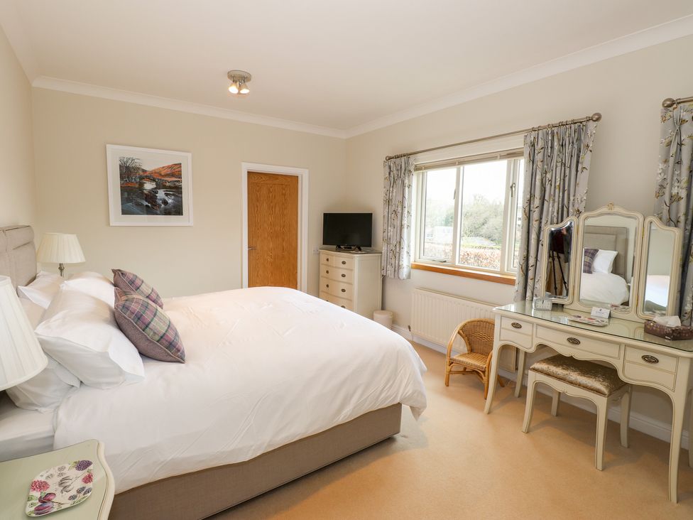 A bedroom with a bed pillows a dresser a TV a vanity table with a mirror and a window with curtains at Edencroft in Greystoke