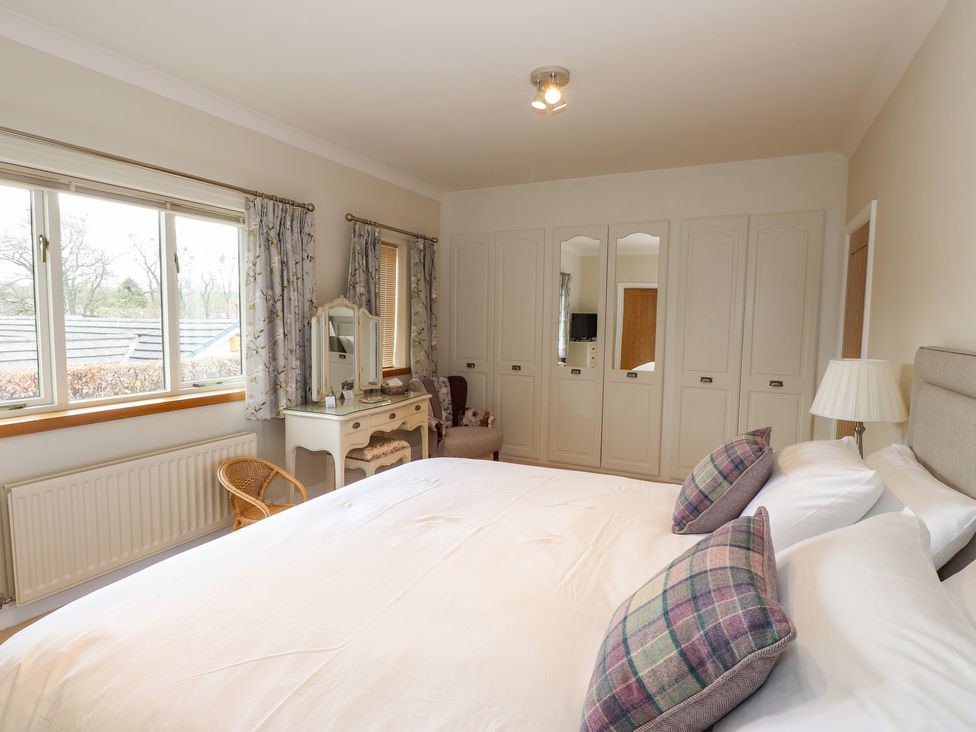A bedroom with a bed with pillows a dressing table armchair wardrobe and windows with curtains at Edencroft in Greystoke