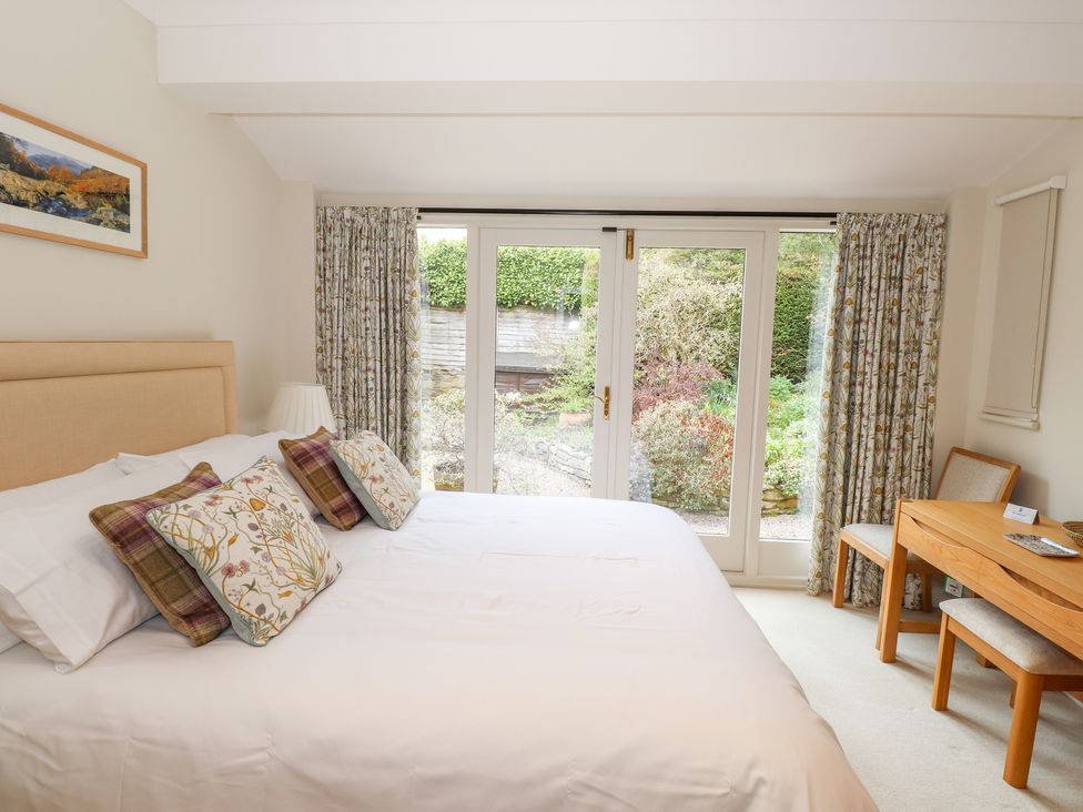 A bedroom with a bed with patterned pillows next to glass doors with curtains and a wooden desk with a chair at Edencroft in Greystoke