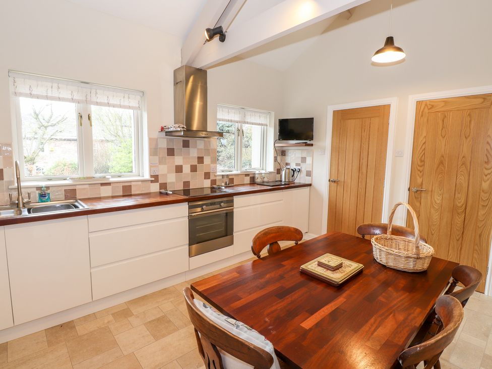 A kitchen with wooden dining table and chairs a wicker basket on the table a countertop with stove sink and windows two wooden doors and a small wall-mounted TV at Edencroft in Greystoke
