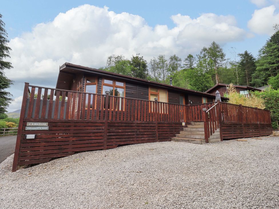 A wooden cabin with a fenced porch and stairs on a gravel area surrounded by trees at Pheasant's Nest Kirkstone 11 Limefitt Park near Windermere