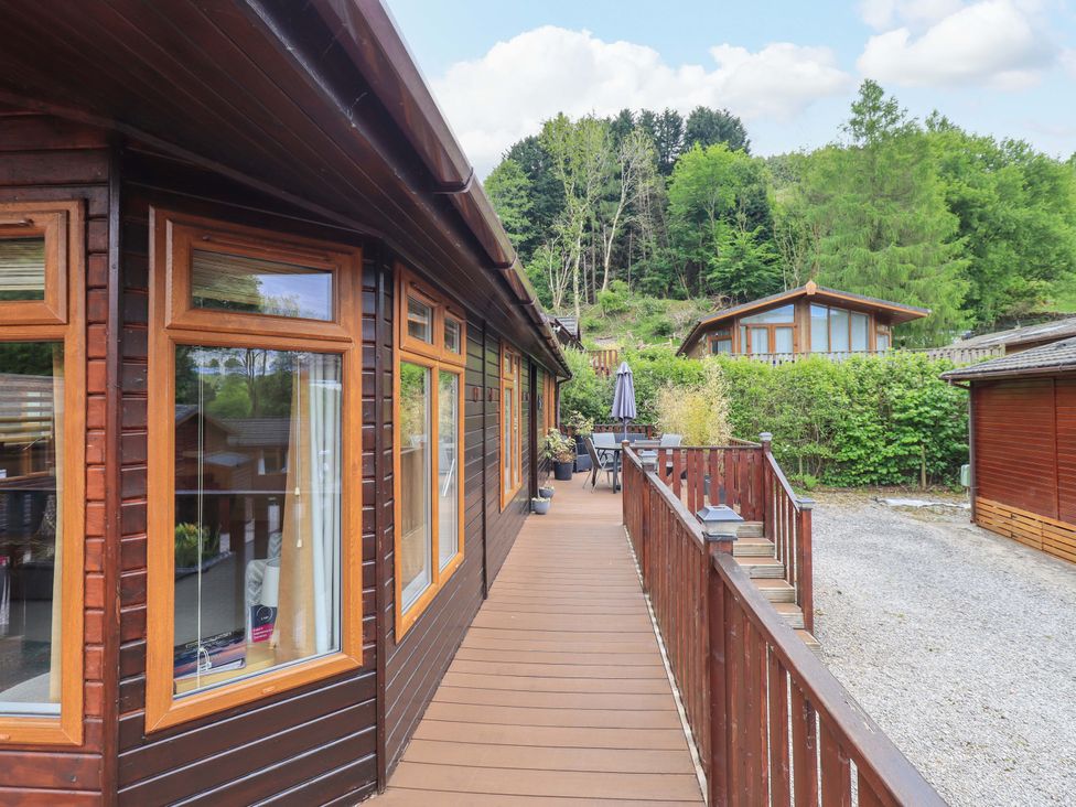 Wooden deck walkway with railings outside wooden cabins surrounded by trees and greenery at Pheasant's Nest Kirkstone 11 Limefitt Park near Windermere