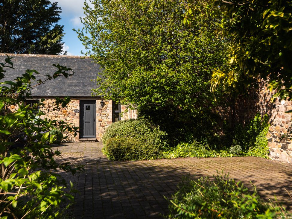A stone building with a door and window surrounded by trees and bushes at Bramble Cottage in Akeld near Wooler