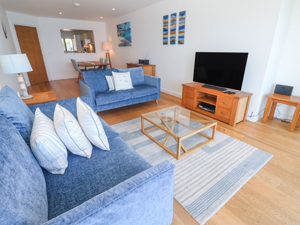 A living room with blue sofas a glass coffee table a wooden TV stand and a dining area at Kittiwake in Carbis Bay