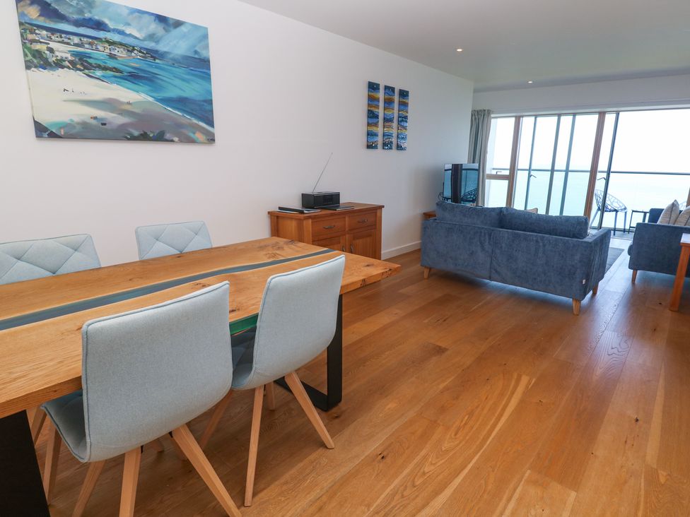 A dining area with wooden table and blue chairs next to a living room with blue sofas and a TV by a balcony with ocean view at Kittiwake in Carbis Bay