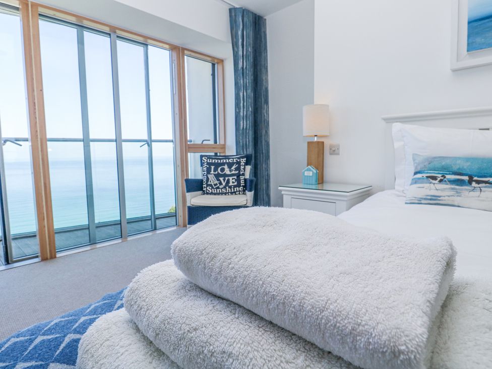 A bedroom with a bed a nightstand and a chair by large glass doors overlooking the sea at Kittiwake in Carbis Bay