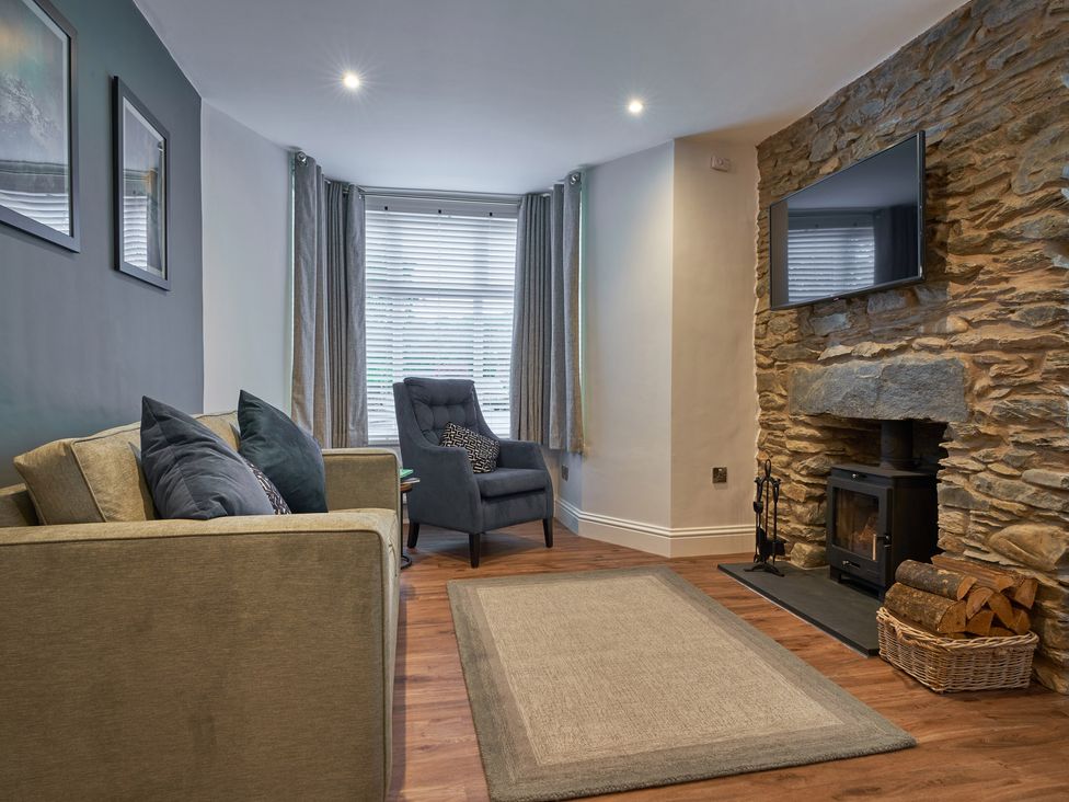 A living room with a sofa, armchair, rug, wood burning stove in a stone fireplace and a wall mounted TV at Lower Sheriff's Place in Windermere