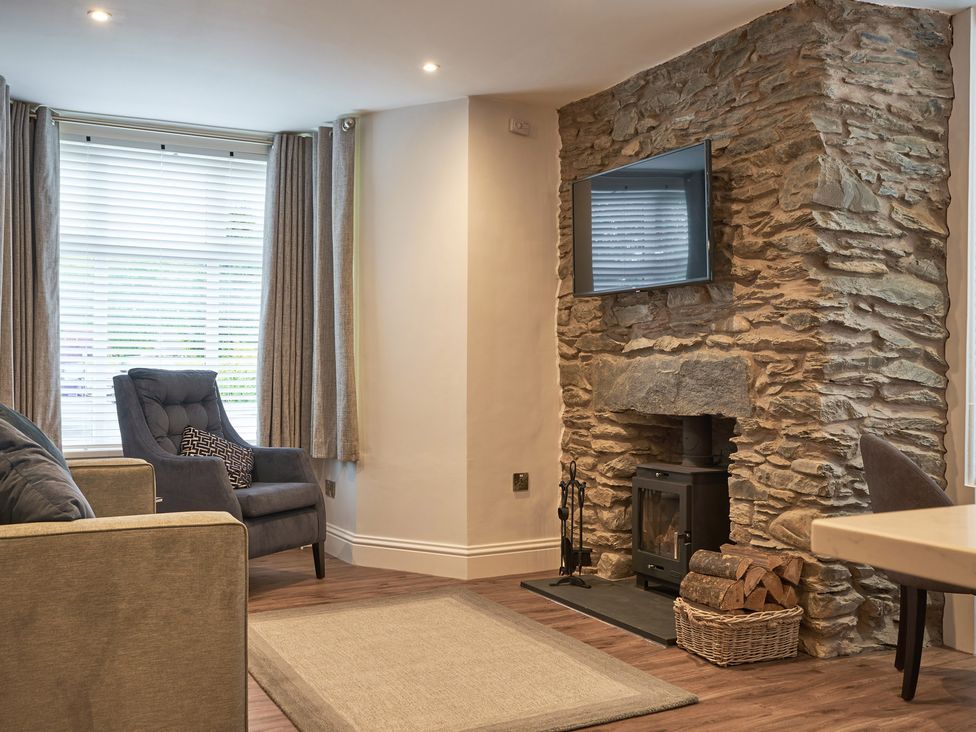 A living room with a stone fireplace wood stove a wall-mounted TV armchair and a window with blinds and curtains at Lower Sheriff's Place in Windermere